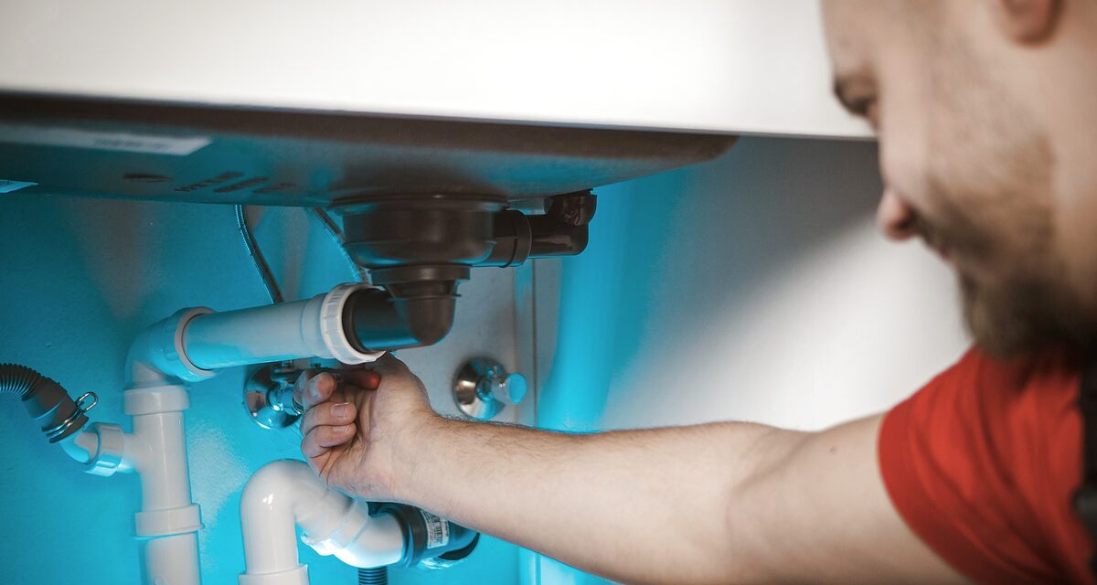 Plumber in a red shirt fixing the P-trap pipes under a kitchen sink